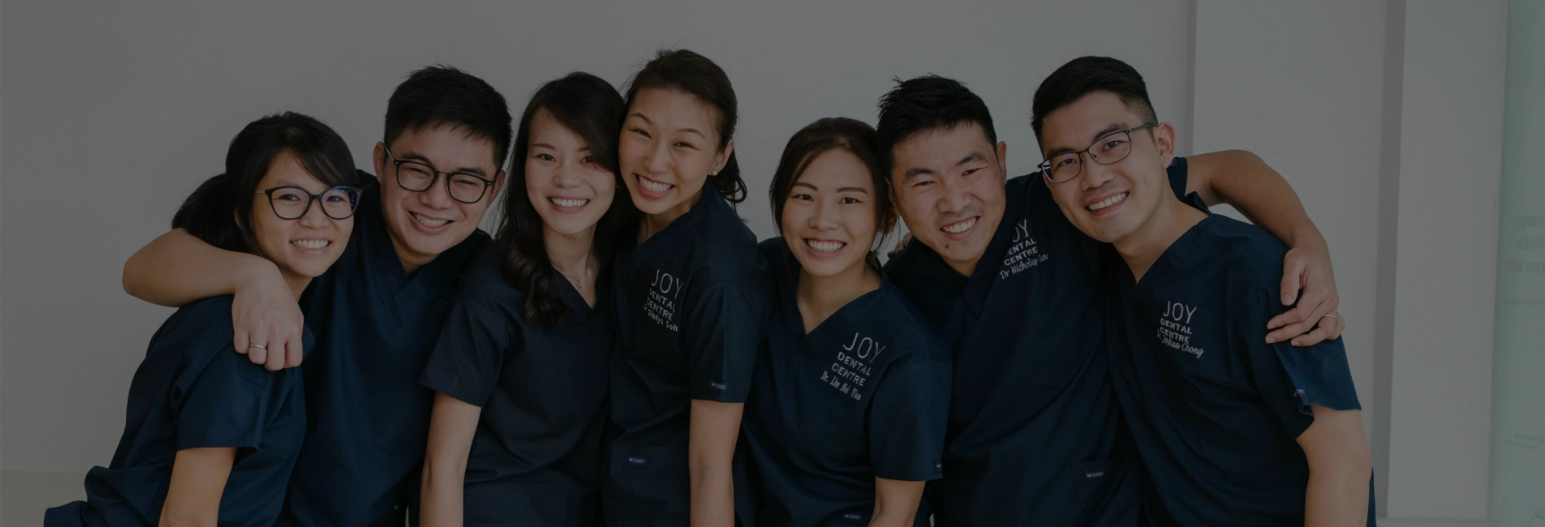 Group portrait: seven Joy Dental Centre staff, smiling in navy uniforms with arms around each other.