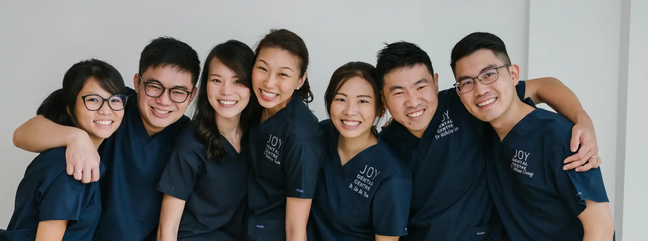 Group portrait: seven Joy Dental Centre staff, smiling in navy uniforms with arms around each other.