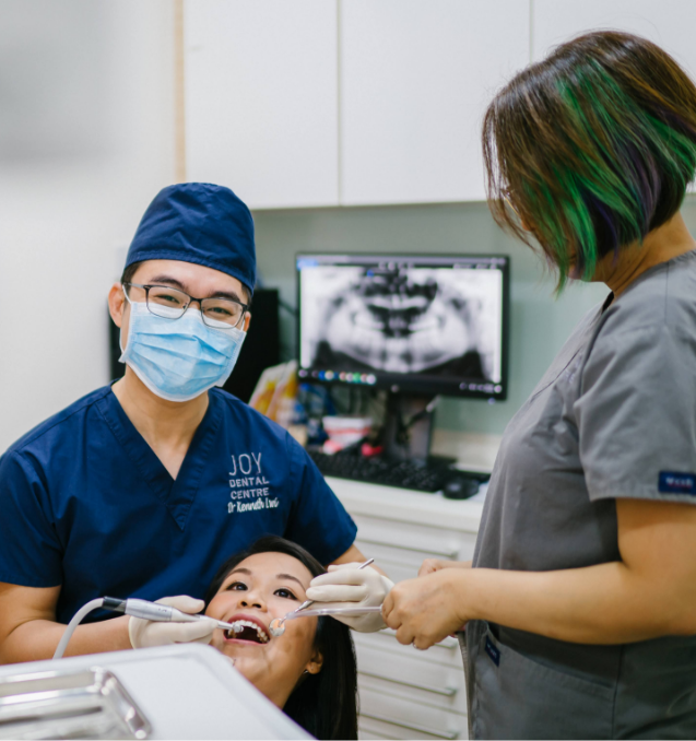 Male dentist in mask and glasses operates on patient lying in dental chair, with a dental assistant handing tools.